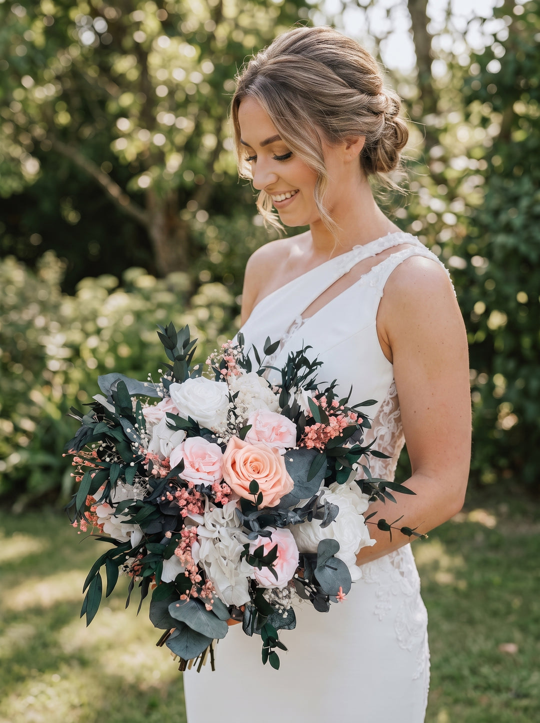 A smiling bride in a white one-shoulder lace wedding gown holds a large bridal bouquet with blush pink roses, white flowers, and green foliage outdoors in a garden setting.