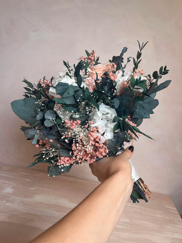 A hand holding a preserved floral bouquet with dusty pink flowers, white blossoms, and green foliage against a soft beige wall and light wood surface.