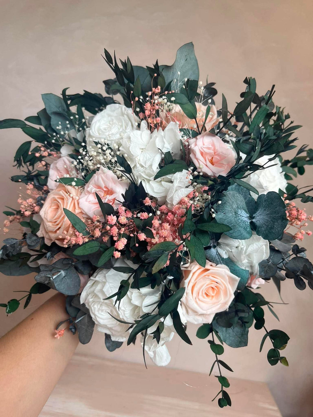 A bridal bouquet featuring white and blush pink roses, green eucalyptus leaves, and delicate baby's breath, held by a person's hand against a soft beige background.