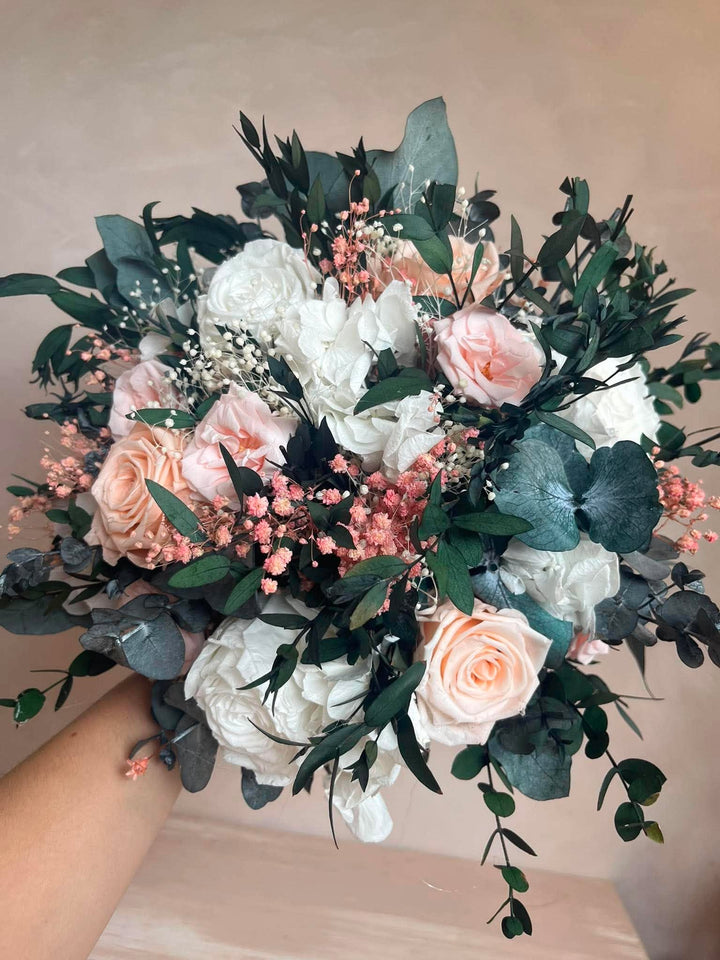 A bridal bouquet featuring white and blush pink roses, green eucalyptus leaves, and delicate baby's breath, held by a person's hand against a soft beige background.