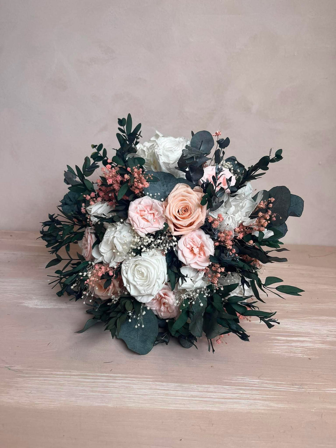 A round floral bouquet featuring white and blush pink roses, dusty pink small flowers, and green eucalyptus leaves, displayed on a light wood surface against a textured beige background.