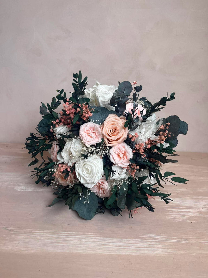 A round floral bouquet featuring white and blush pink roses, dusty pink small flowers, and green eucalyptus leaves, displayed on a light wood surface against a textured beige background.