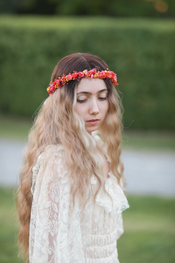 A woman with long wavy blonde hair wearing a red and orange floral crown and a cream lace top, standing outdoors against a blurred green hedge background.