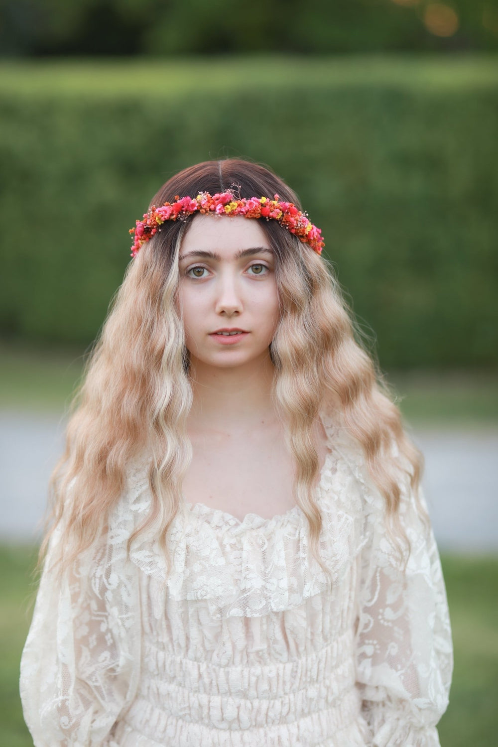 A young woman with long wavy blonde hair wearing a red and orange floral crown and a cream lace dress, standing outdoors against a blurred green hedge background.