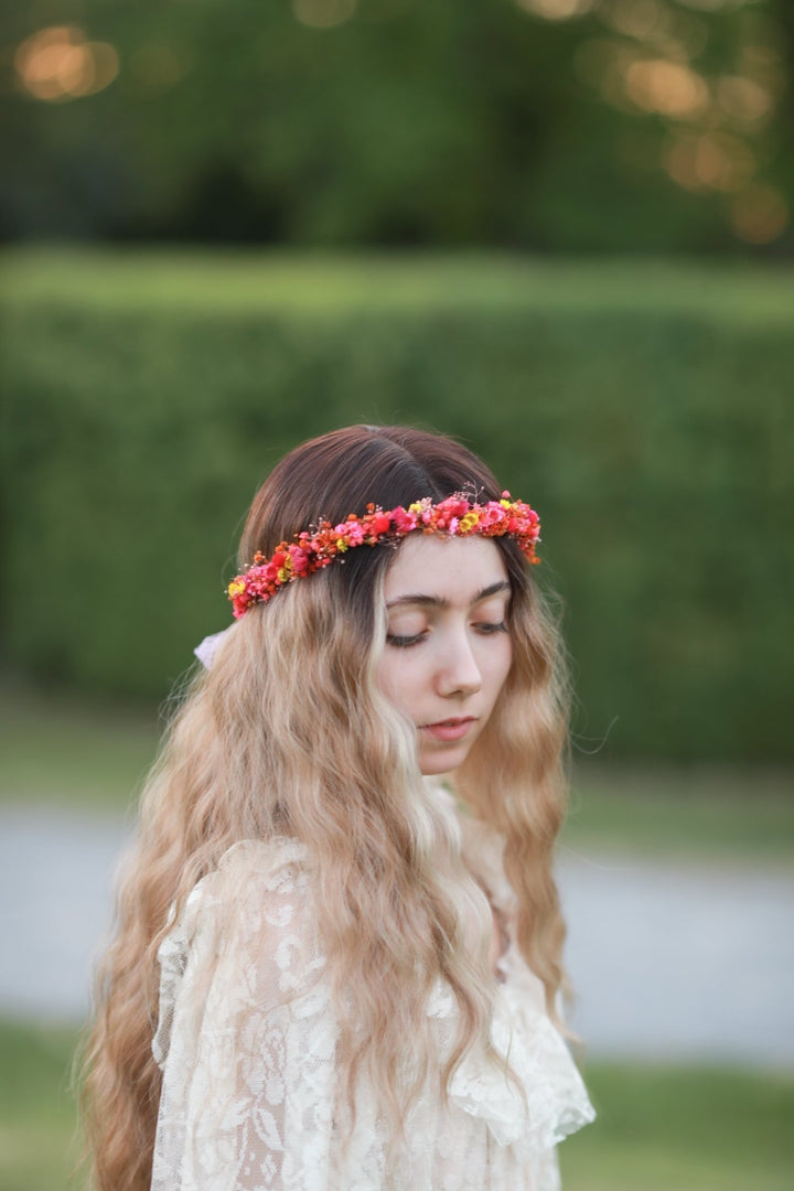 A woman outdoors wearing a red and orange floral crown and a cream lace top, with a blurred green garden background.