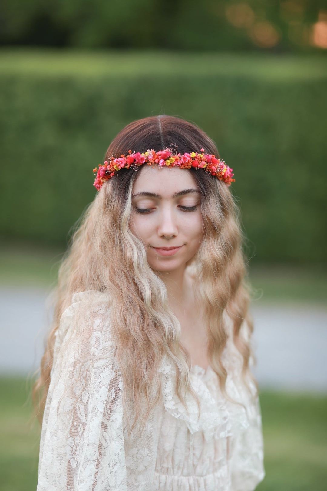 A woman outdoors wearing a floral crown of pink, red, and yellow flowers, with long wavy blonde hair and a cream lace top.