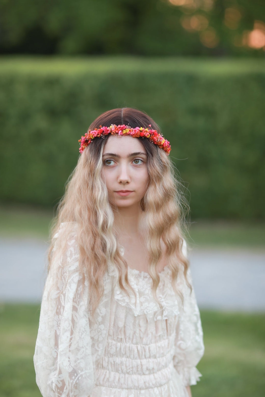 A young woman with long wavy blonde hair wearing a red and orange floral crown and a cream lace dress, standing outdoors in front of a blurred green hedge.