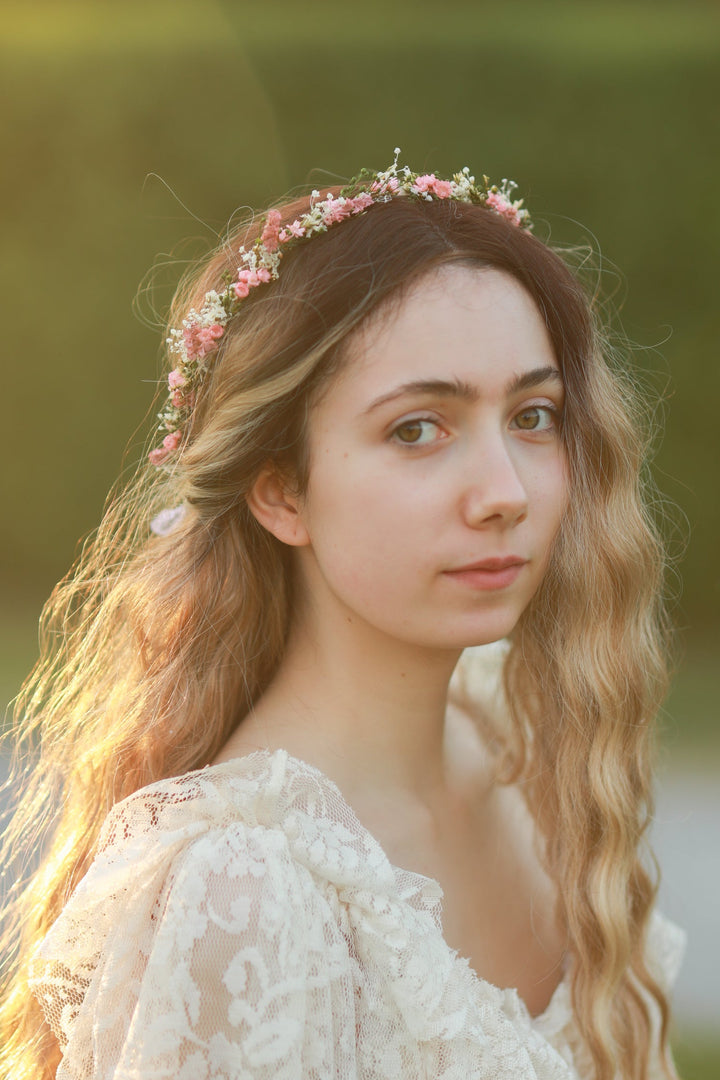 A young woman with long wavy light brown hair wearing a delicate pink and white floral crown, looking off-camera with a soft, dreamy expression against a blurred green and gold outdoor background.