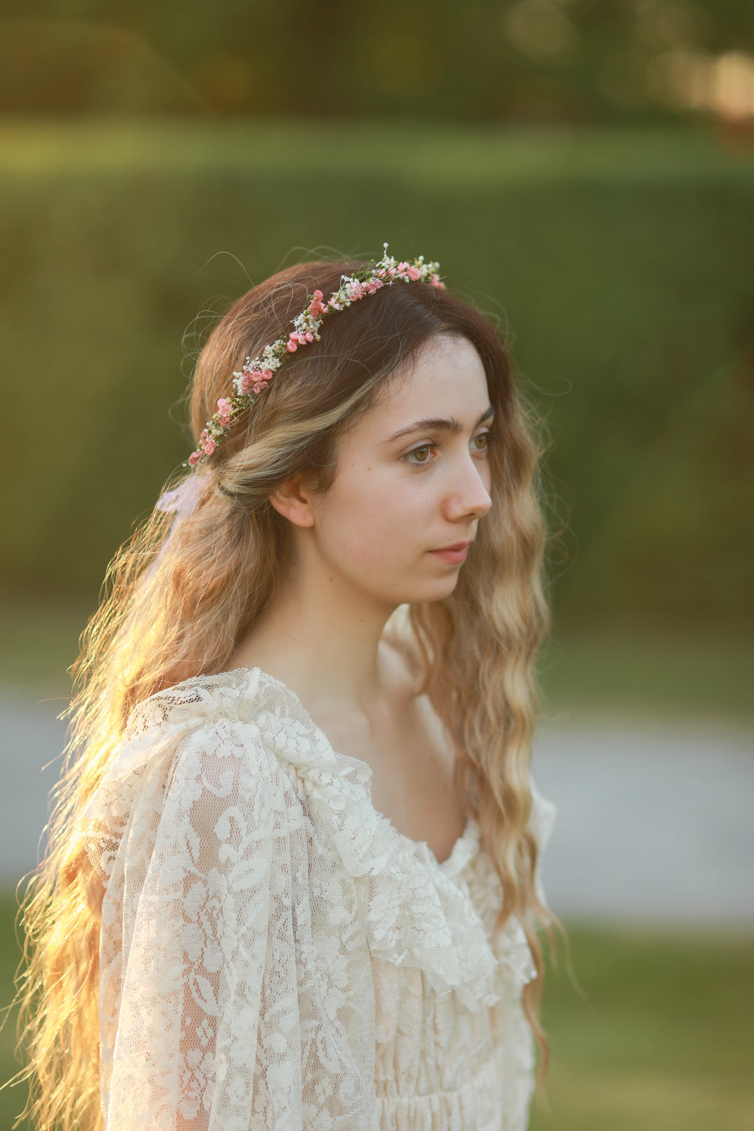 A woman in profile wearing a delicate floral crown and a cream lace dress, photographed outdoors with a soft blurred green background.