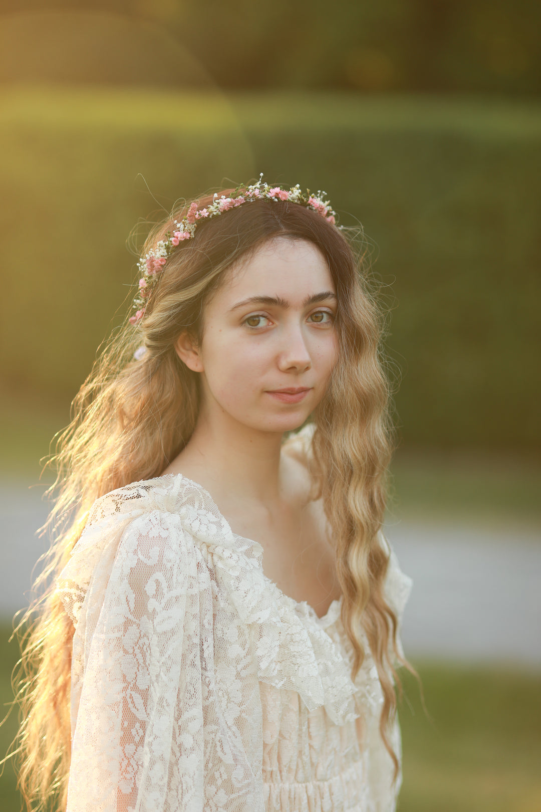 A young woman with long wavy light brown hair wearing a delicate pink and white floral crown and a white lace boho-style dress, photographed outdoors in warm golden hour sunlight with a blurred green background.