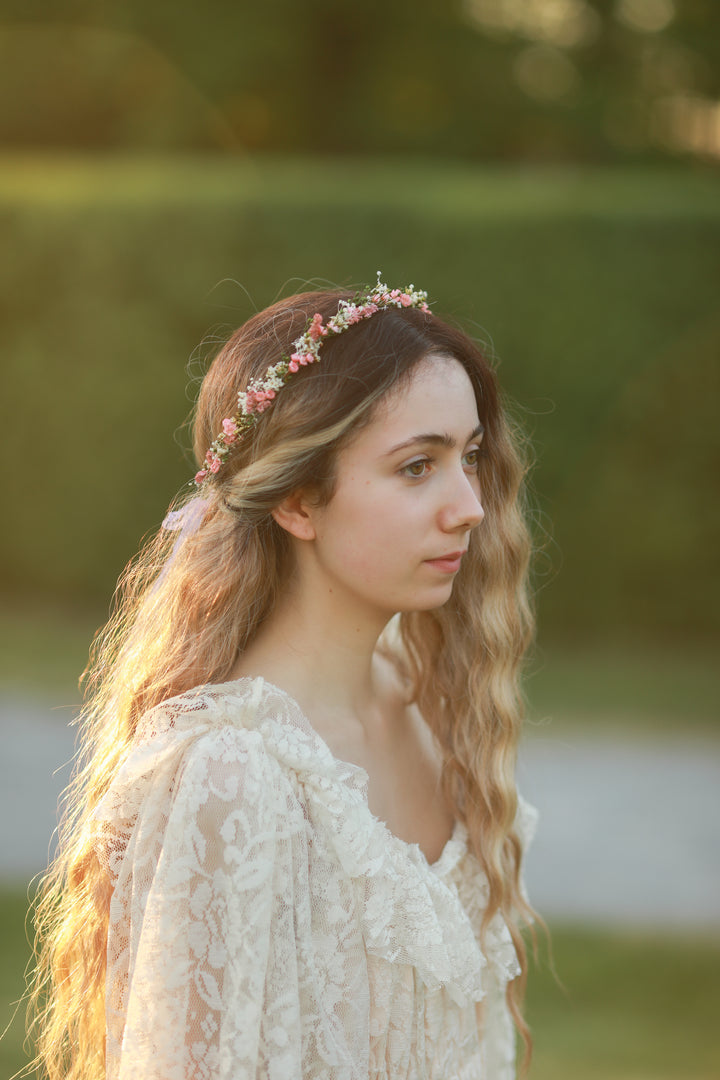 A woman in profile wearing a delicate floral crown and a cream lace top, photographed outdoors in warm golden-hour light against a blurred green background.