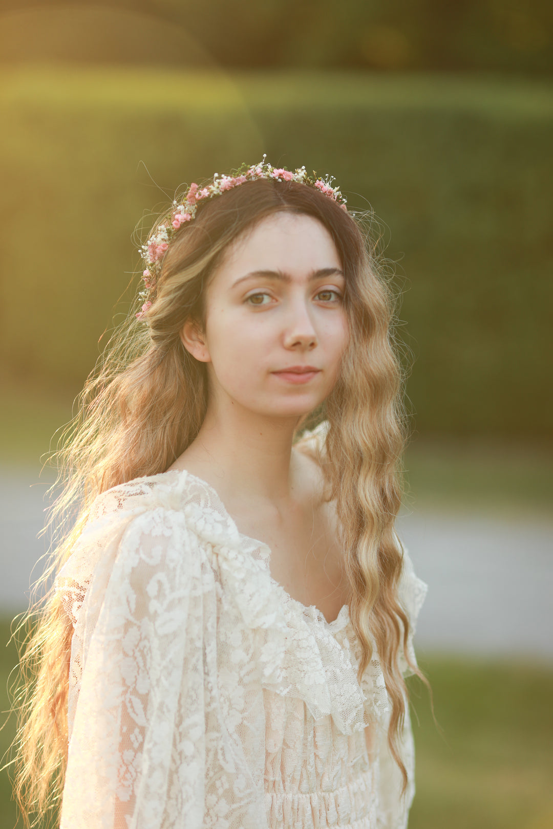 A young woman with long wavy light brown hair wearing a delicate pink floral crown and a cream lace boho-style dress, photographed outdoors in warm golden hour sunlight with a blurred green background.
