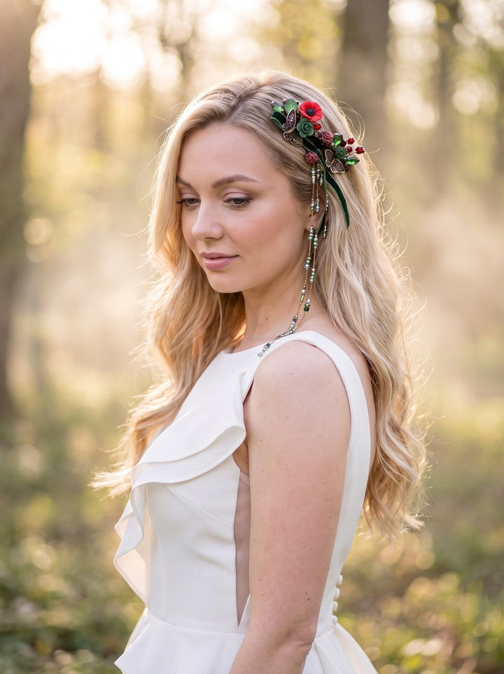A woman in a white sleeveless dress wearing a floral headpiece with red flowers and green stones, standing in a sunlit forest setting.