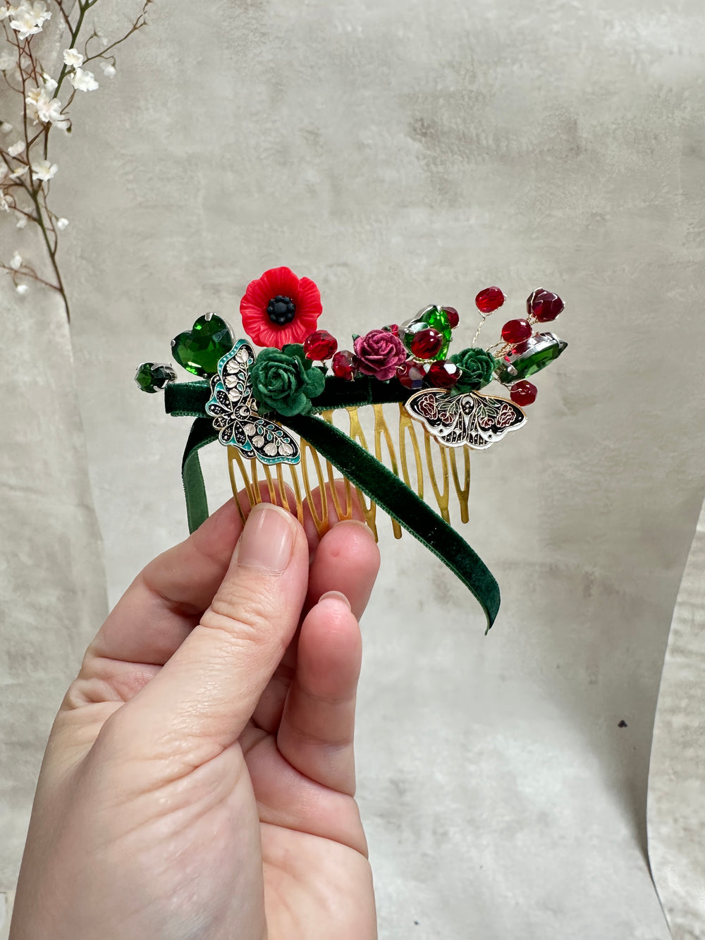A hand holds a decorative hair comb featuring green velvet leaves, red and pink fabric flowers, green glass beads, and silver butterfly embellishments against a neutral textured background.