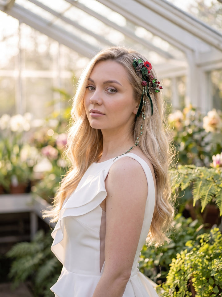 A blonde woman in a white ruffled sleeveless dress stands in a sunlit greenhouse, wearing a floral hair accessory with red flowers and green beads.