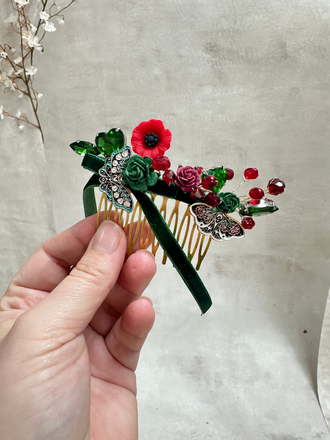 A hand holding a decorative hair comb featuring green and red flowers, green leaves, and silver butterfly embellishments, set against a textured light gray background.