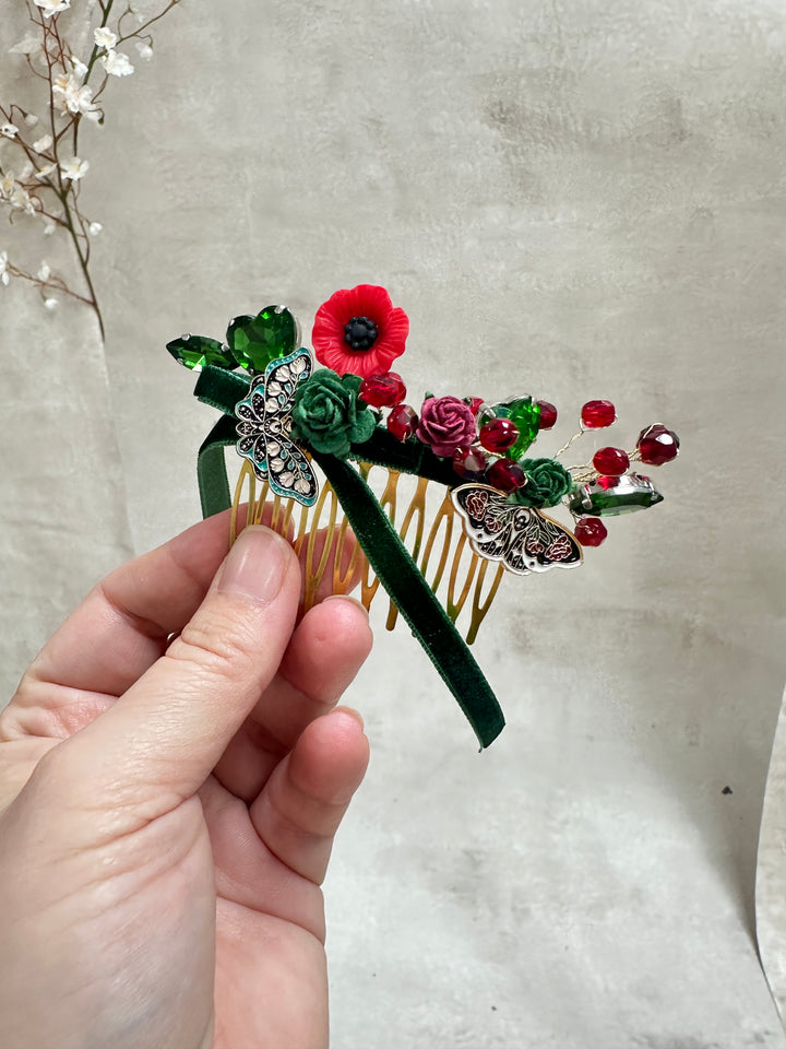 A hand holding a decorative hair comb featuring green and red flowers, green leaves, and silver butterfly embellishments, set against a textured light gray background.