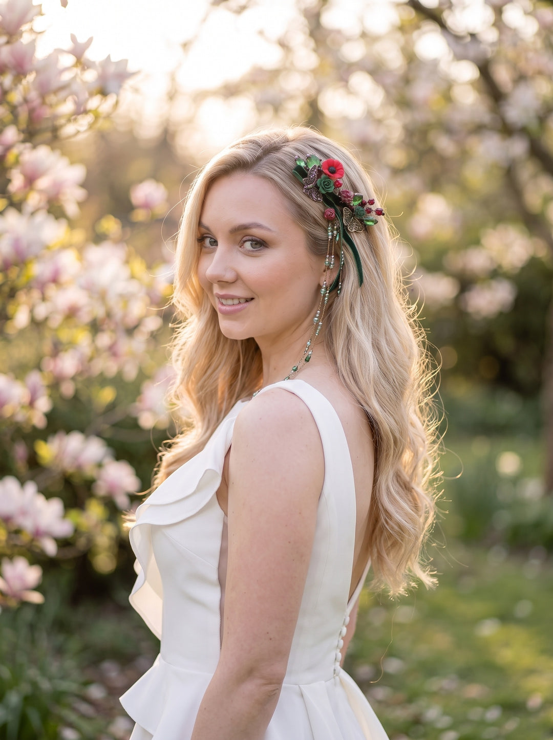 A woman outdoors wearing a sleeveless white dress and a floral headpiece with red and green flowers, photographed in a sunlit garden with blurred pink blossoms in the background.