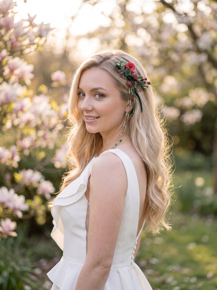 A woman outdoors wearing a sleeveless white dress and a floral headpiece with red and green flowers, photographed in a sunlit garden with blurred pink blossoms in the background.