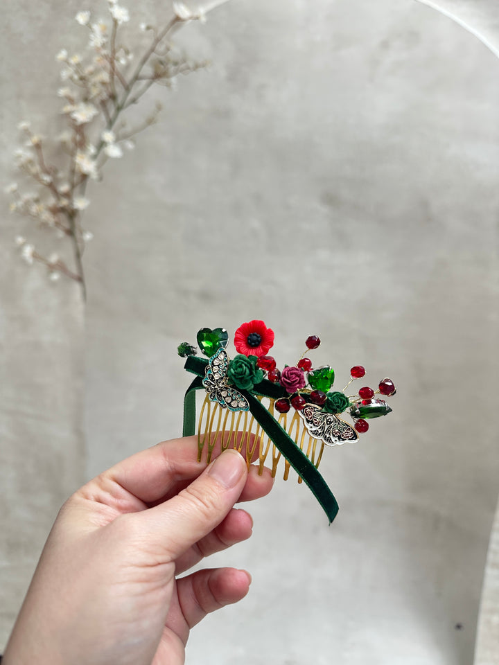 A hand holding a decorative hair comb with a green butterfly and floral design featuring red and green rhinestones on a light gray background.