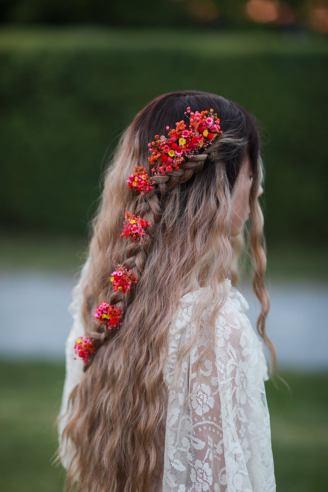 A young girl with long, wavy blonde-highlighted hair is shown from the back outdoors. Her hair features a braided section adorned with bright red and orange flower-like hair clips, and she is wearing a white lace top.