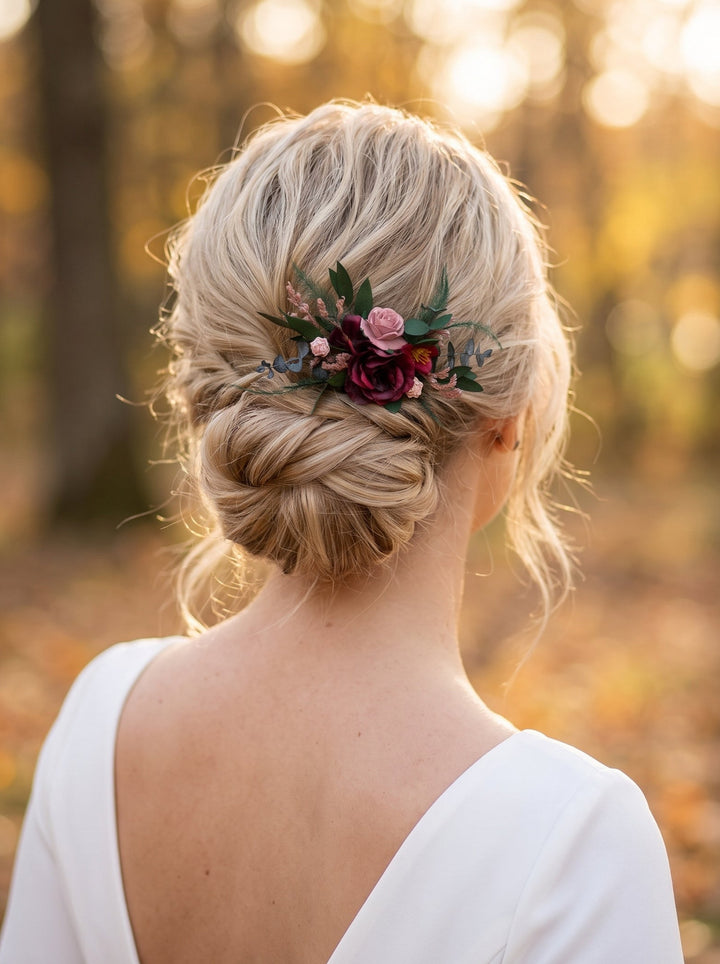 A blonde bride viewed from behind, wearing a white wedding dress and a braided updo decorated with a floral hair accessory featuring burgundy and pink roses with green leaves, set against a warm, blurred autumn forest background.