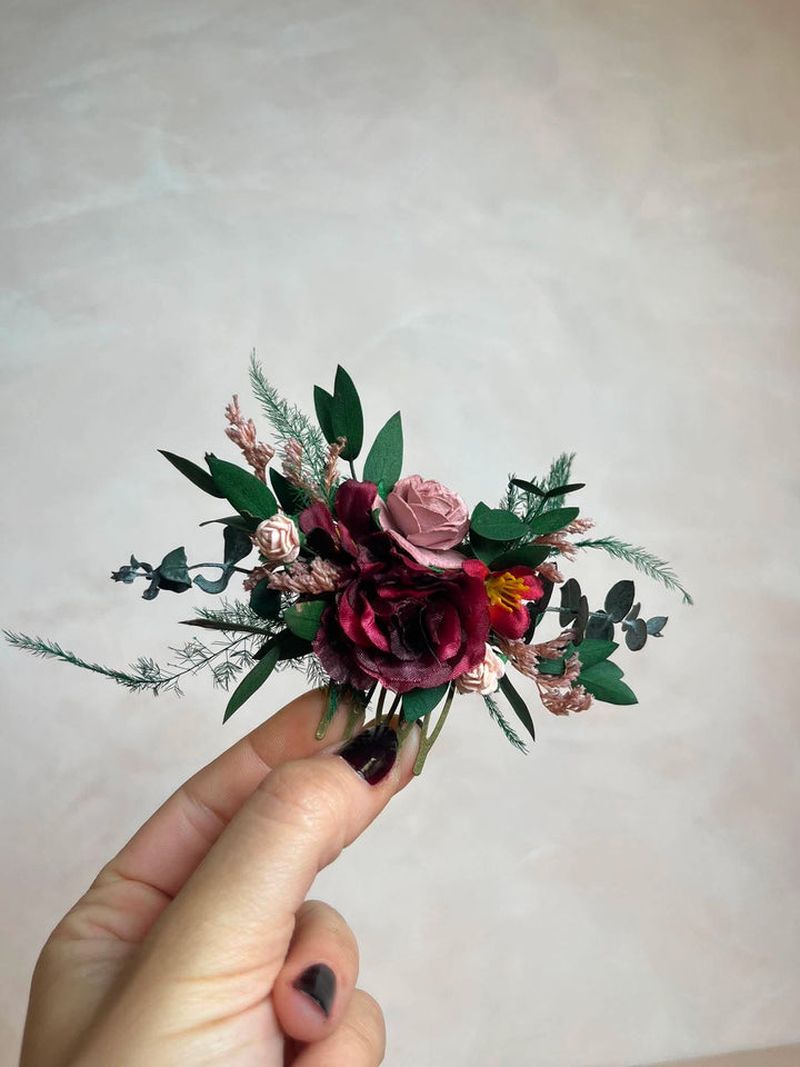 A burgundy bridal hair comb  held by a hand, featuring burgundy and dusty pink roses, green leaves, against a neutral beige background.