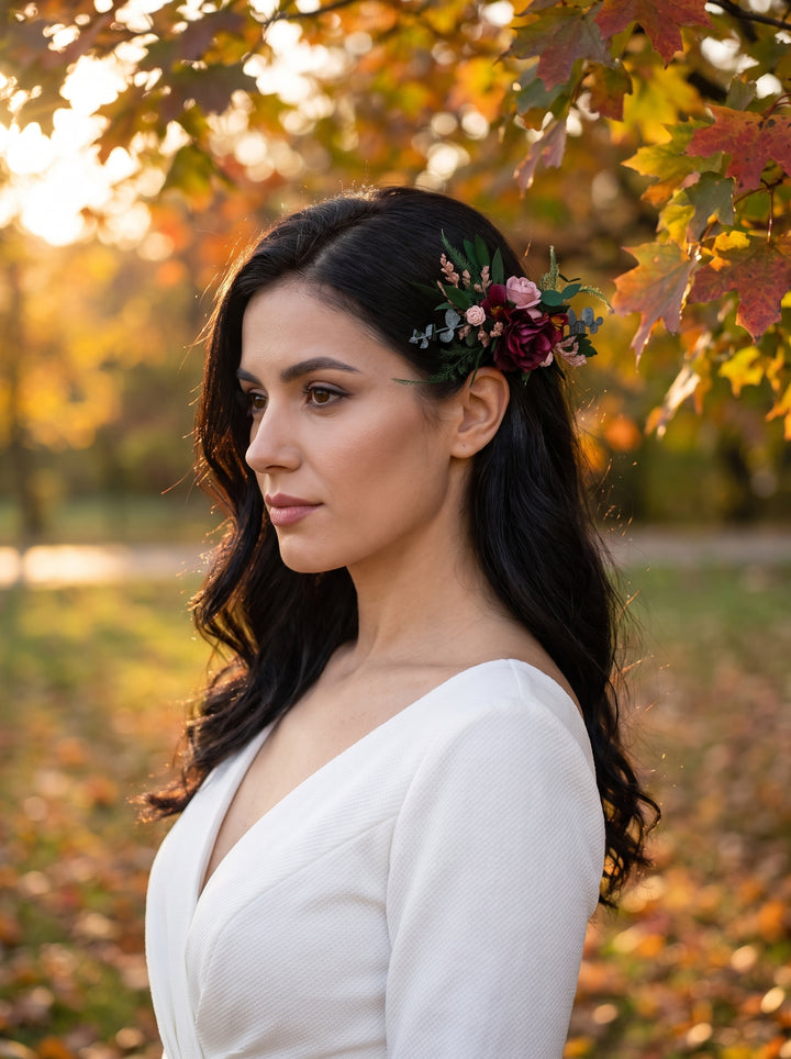 A woman in a white V-neck top wearing a floral hair accessory with burgundy, pink, and green flowers, photographed outdoors in a warm autumn park setting.