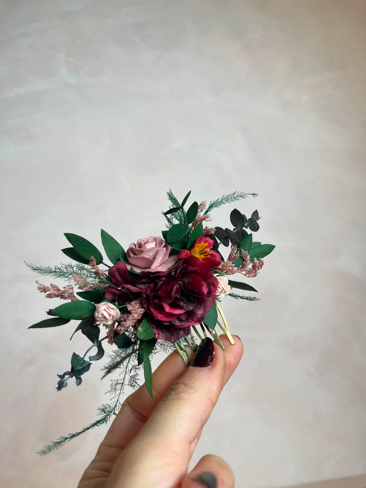 A hand with dark nail polish holds a small floral hair comb  featuring burgundy and pink roses, green leaves, and delicate filler stems against a soft beige background.