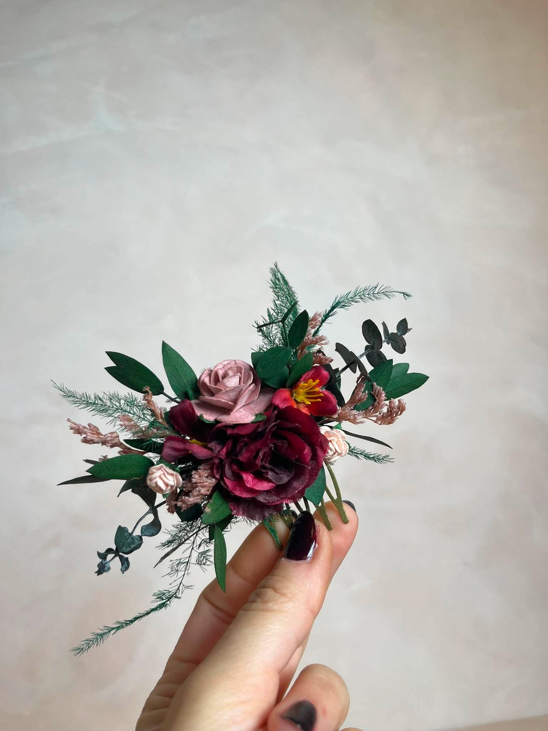 A hand with dark nail polish holds a small hair comb  featuring burgundy and dusty pink roses, green foliage, and a small yellow accent flower against a neutral beige background.