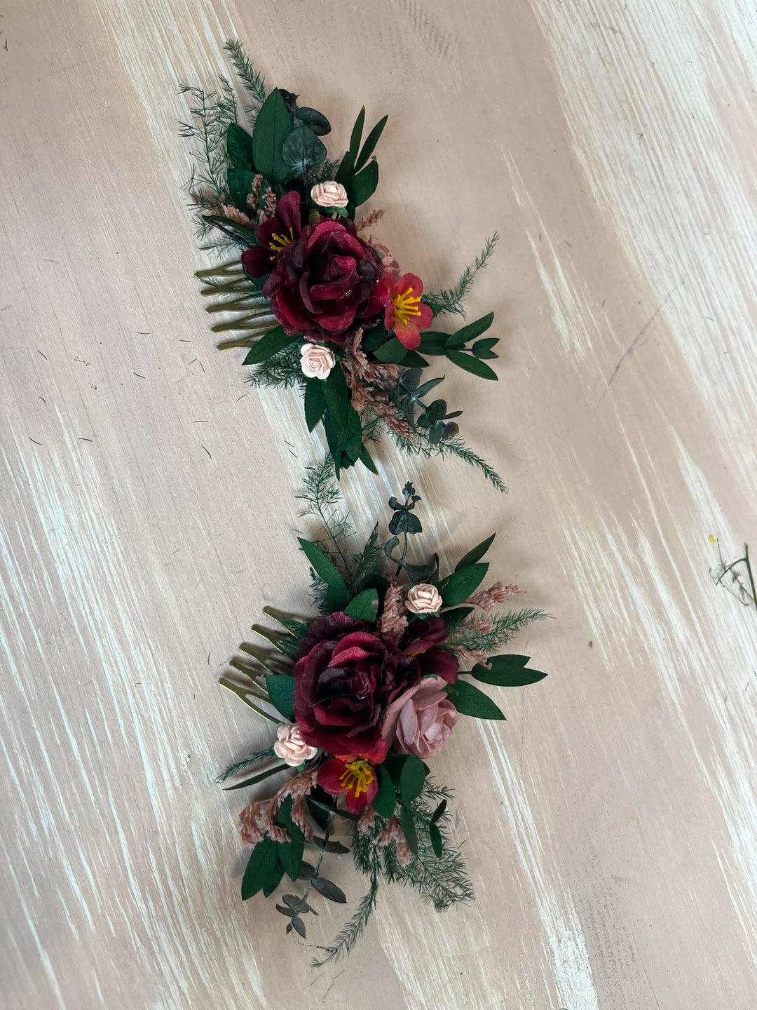 Two matching floral hair combs with burgundy roses, green foliage, and small pink and yellow accents, arranged on a light wood background.