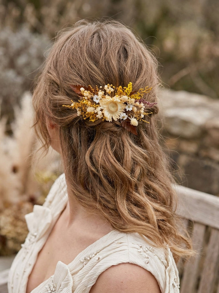 A woman shown from the back wearing a bohemian-style dried flower hair accessory in her hair, featuring a cream flower center with yellow, white, and brown petals, set against a soft-focus outdoor background.
