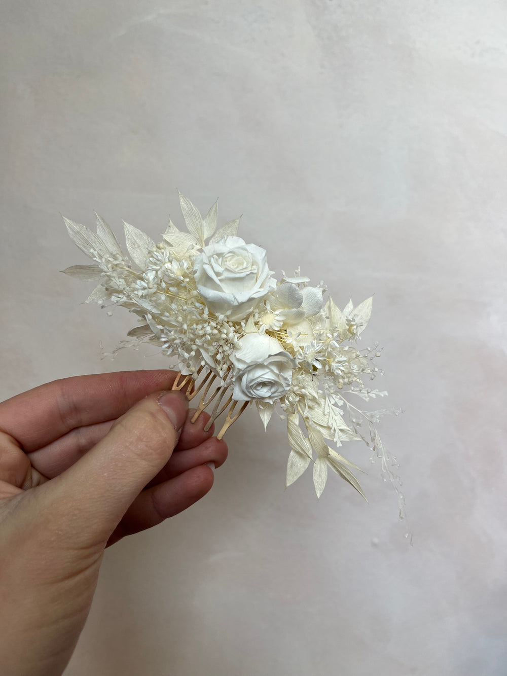 A hand holding a dried floral hair comb featuring white and cream roses, delicate leaves, and small filler flowers on a light beige background.