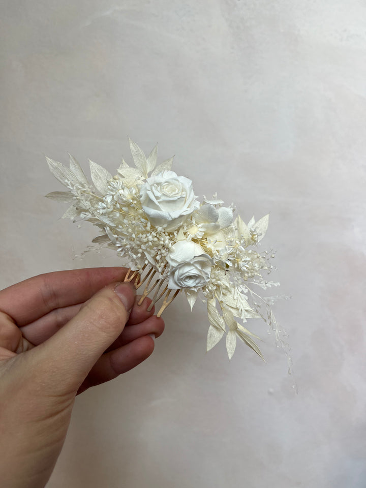 A hand holding a dried floral hair comb featuring white and cream roses, delicate leaves, and small filler flowers on a light beige background.