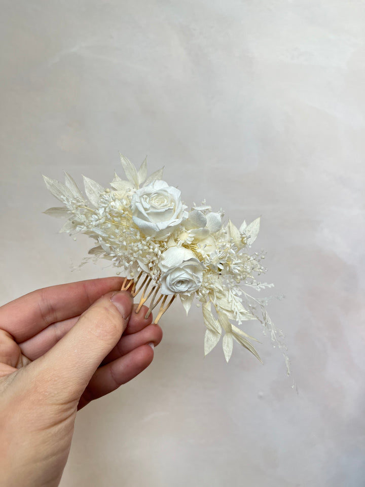 A hand holding a small dried floral hair comb accessory with white and cream roses, delicate leaves, and fine stems against a soft light gray background.