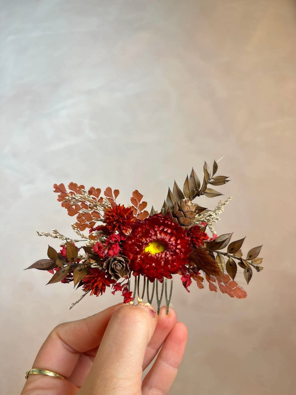 Hair comb crafted entirely from preserved and dried botanicals in deep, rich autumn tones - vivid crimson strawflowers, velvety dried roses, rustic pine cones, warm brown leaves, and delicate dried grasses.