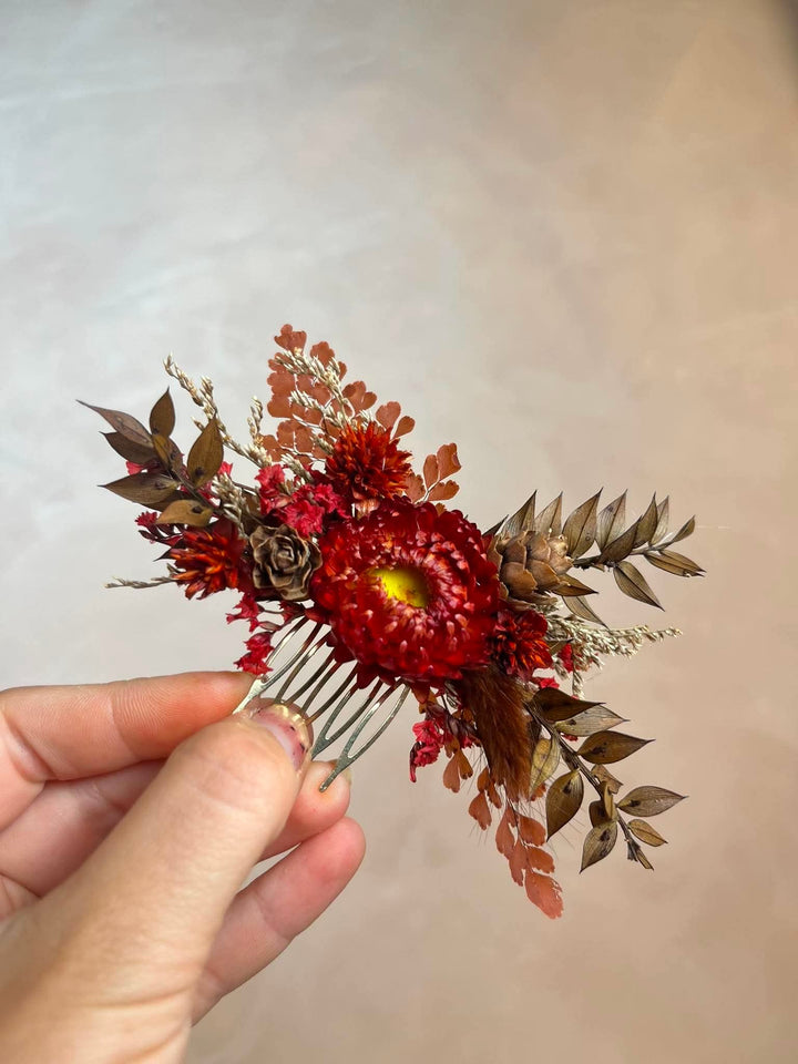 Hair comb crafted entirely from preserved and dried botanicals in deep, rich autumn tones - vivid crimson strawflowers, velvety dried roses, rustic pine cones, warm brown leaves, and delicate dried grasses.