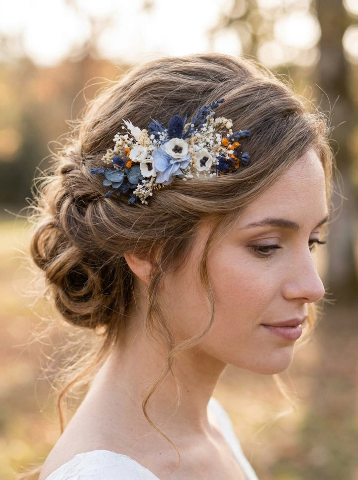 A woman in profile wearing a blue and white floral hair accessory with dried flowers and small amber accents, photographed outdoors against a softly blurred warm-toned background.