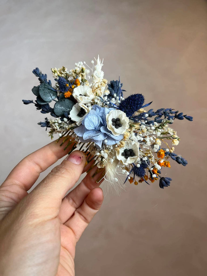 A hand holding a floral hair comb featuring light blue, navy, white, and orange botanical elements against a soft beige background.