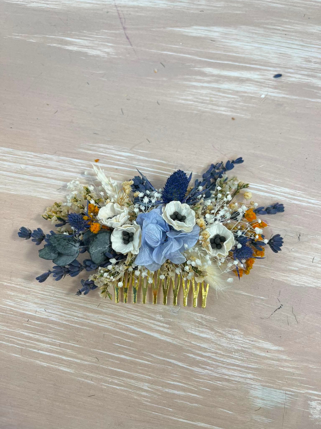 A floral hair comb featuring white anemone flowers, a light blue fabric rose, navy blue lavender, and small orange blossoms, set in a gold metal comb on a distressed beige wooden background.