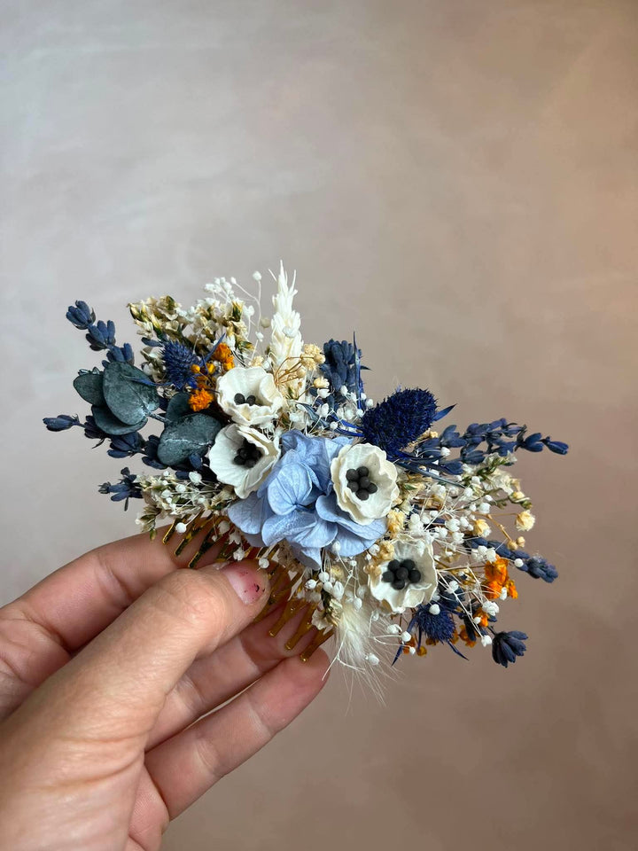 A floral hair accessory held in a hand, featuring light blue and white flowers, navy blue accents, and small orange details against a plain beige background.