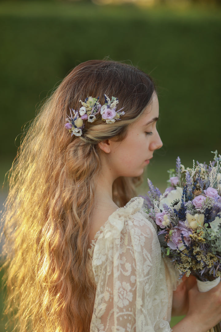 Woman with floral hairpiece and bouquet, standing outdoors with greenery in the background