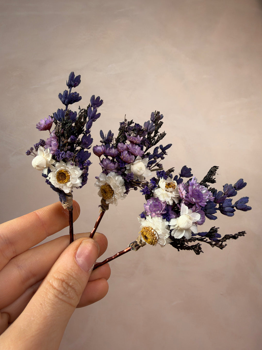 A hand holding three small dried flower bobby pins with purple and white blossoms against a plain beige background.