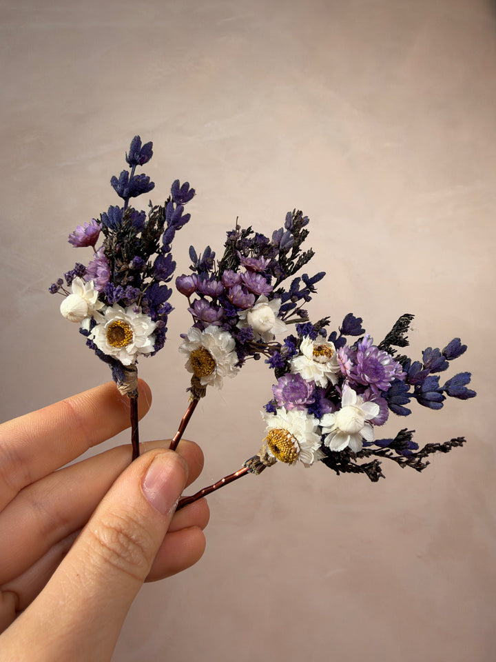 A hand holding three small dried flower bobby pins with purple and white blossoms against a plain beige background.