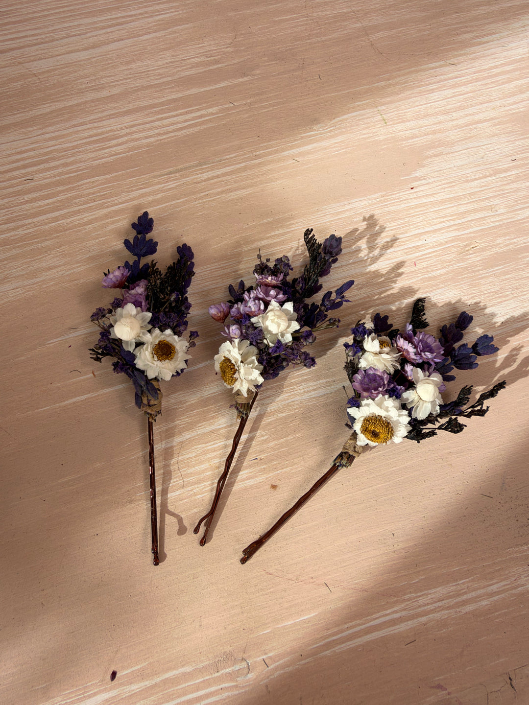 Three small dried flower hair pins with white and purple flowers on a light wood background.