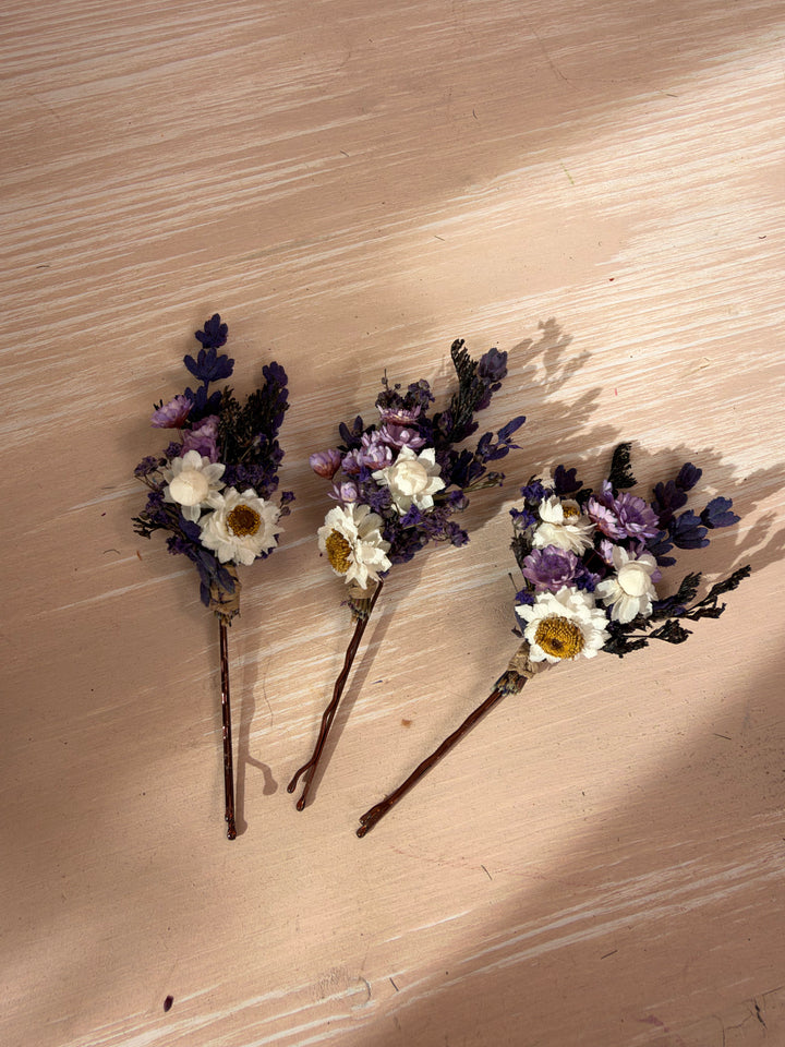 Three small dried flower hair pins with white and purple flowers on a light wood background.