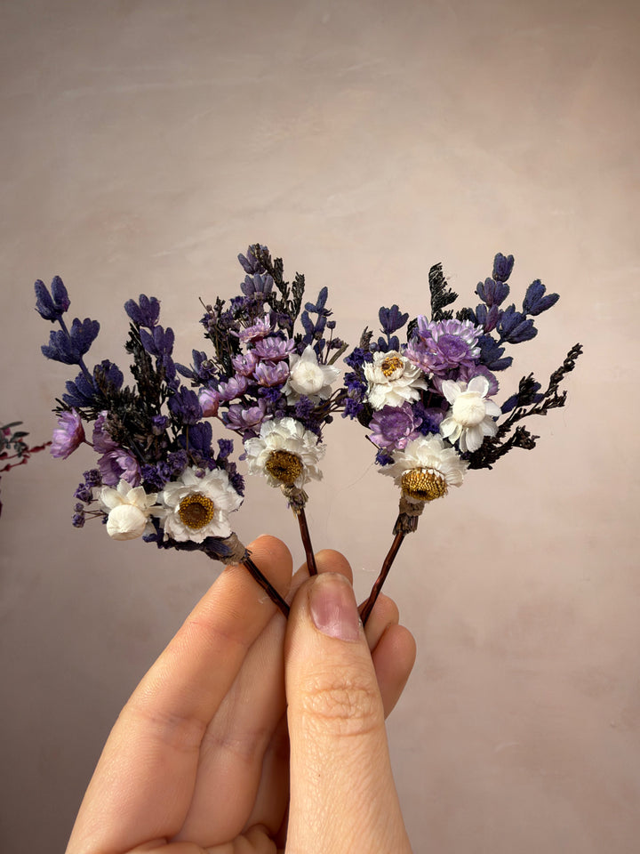 A hand holding two small dried flower bobby pins with purple, lavender, white, and dark purple blossoms against a plain beige background.