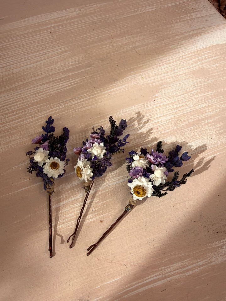 Three small dried flower bobby pins with white and purple flowers on brown stems, arranged on a light wood surface.