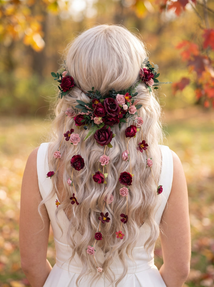 A woman viewed from behind wearing a long, wavy platinum blonde wig adorned with dark red and pink flowers and green leaves, styled with a floral crown in an outdoor autumn setting.
