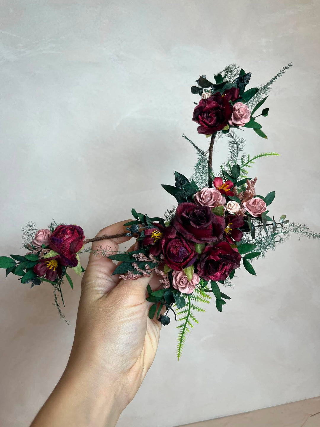 A hand holding a decorative floral arrangement featuring dark burgundy and pink faux roses with green and foliage, set against a plain light beige wall.